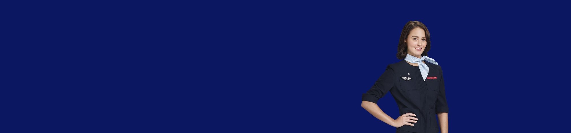 Azafata sonriente, manos en cintura, fondo azul liso, viste uniforme azul oscuro con pañuelo.