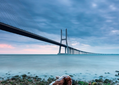 Un puente colgante se extiende sobre agua tranquila, bajo un cielo nublado al amanecer.