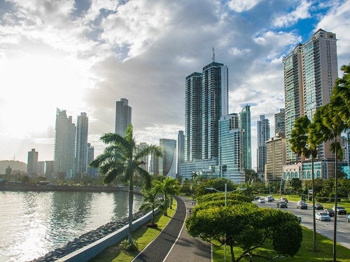 Rascacielos reflejan luz solar mientras bordean un camino junto al mar con palmeras.