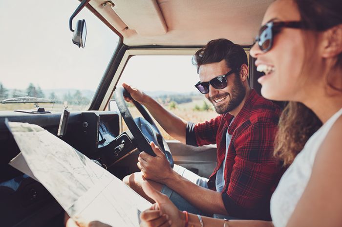 Pareja conduce sonriente en un coche, usando mapa, rodeados de paisaje natural.
