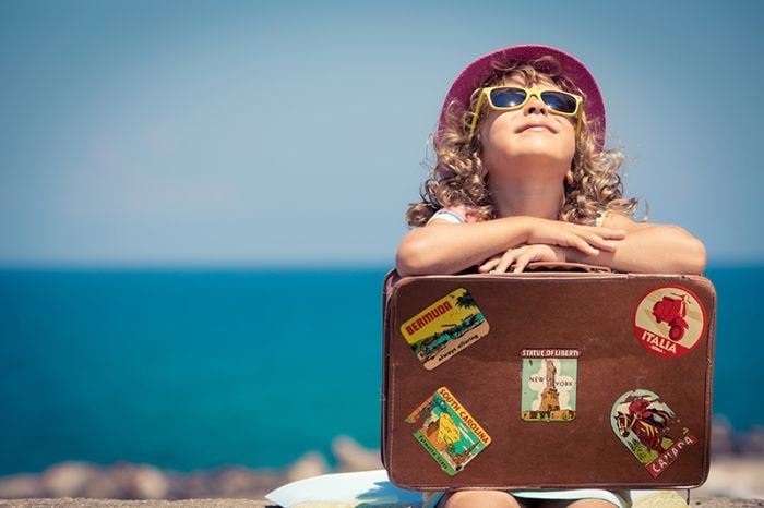 Niño con sombrero y gafas, sonríe apoyado en maleta frente al mar.