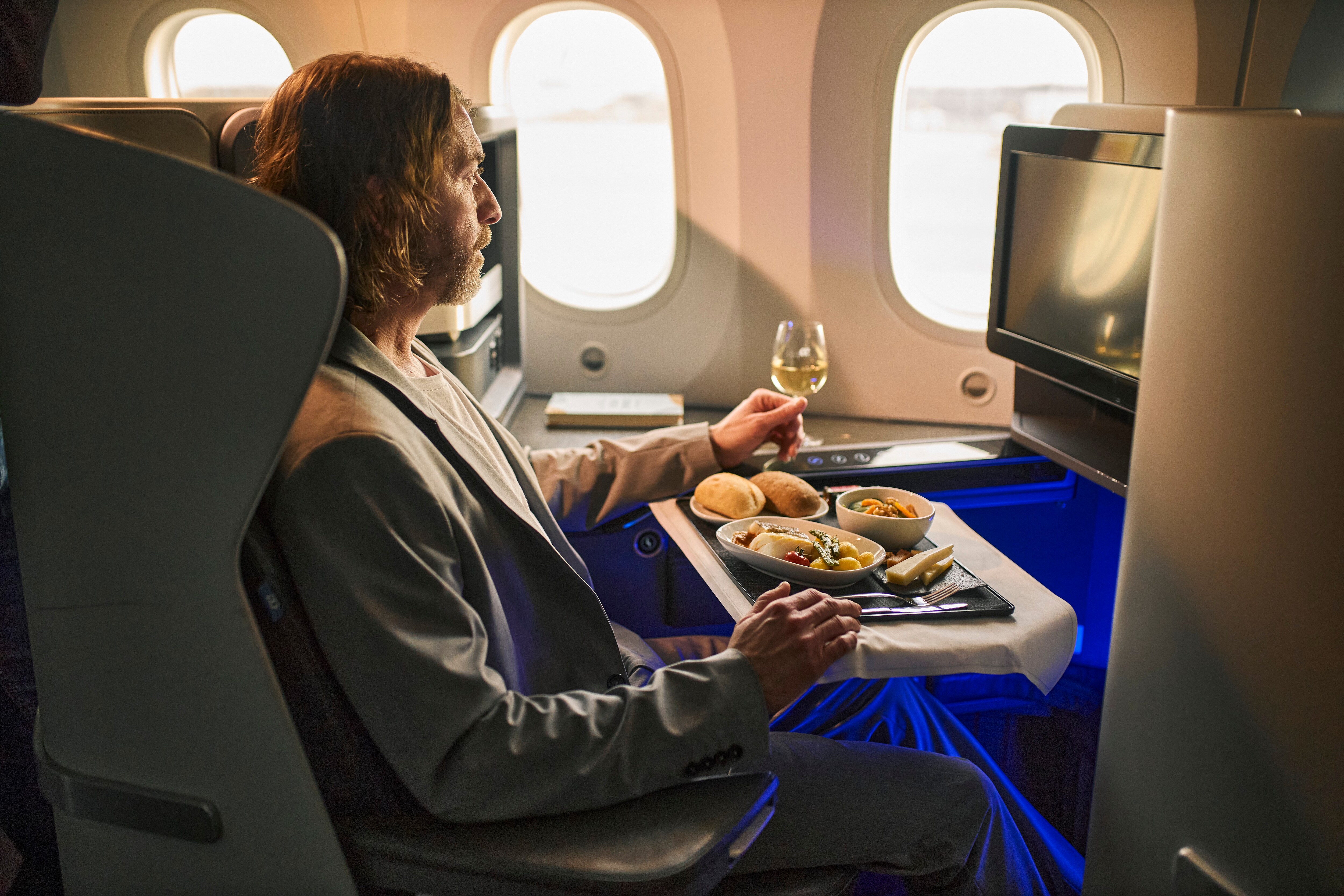 Un uomo è seduto su un aereo e tiene in mano un bicchiere di vino davanti a un vassoio di cibo.